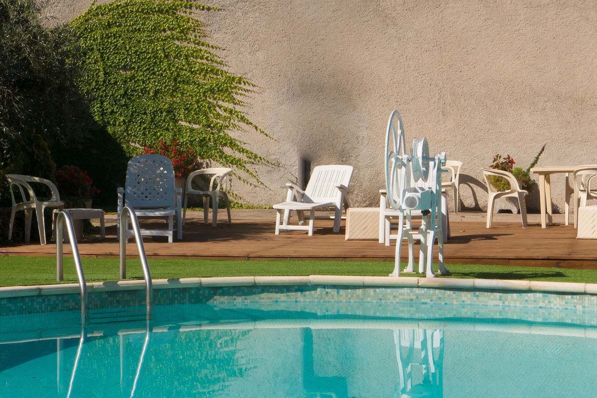 The spacious pool area features a clear blue swimming pool surrounded by a wooden deck. Several white lounge chairs and tables provide seating options, with lush greenery in the background enhancing the tranquil setting.