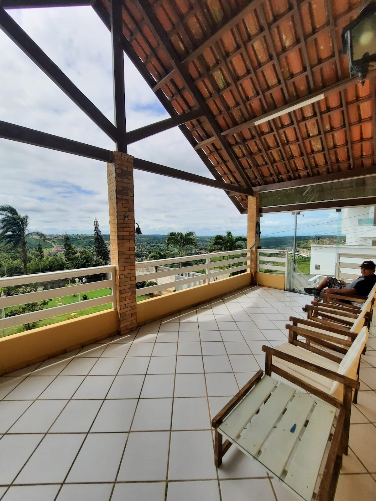 A spacious balcony features wooden chairs arranged for relaxation, with a panoramic view of the surrounding landscape. The sloped roof, supported by wooden beams, complements the open area, creating a comfortable seating environment. Lush greenery and distant hills enhance the natural scenery.