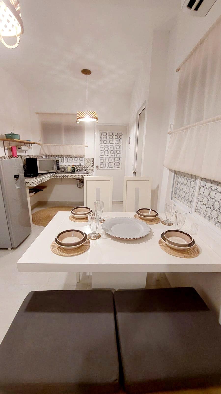 A modern kitchen dining area is presented, featuring a white table set with plates and glasses. A cozy seating arrangement is on one side, while the kitchen, equipped with appliances, is visible in the background. Soft lighting enhances the overall ambiance.