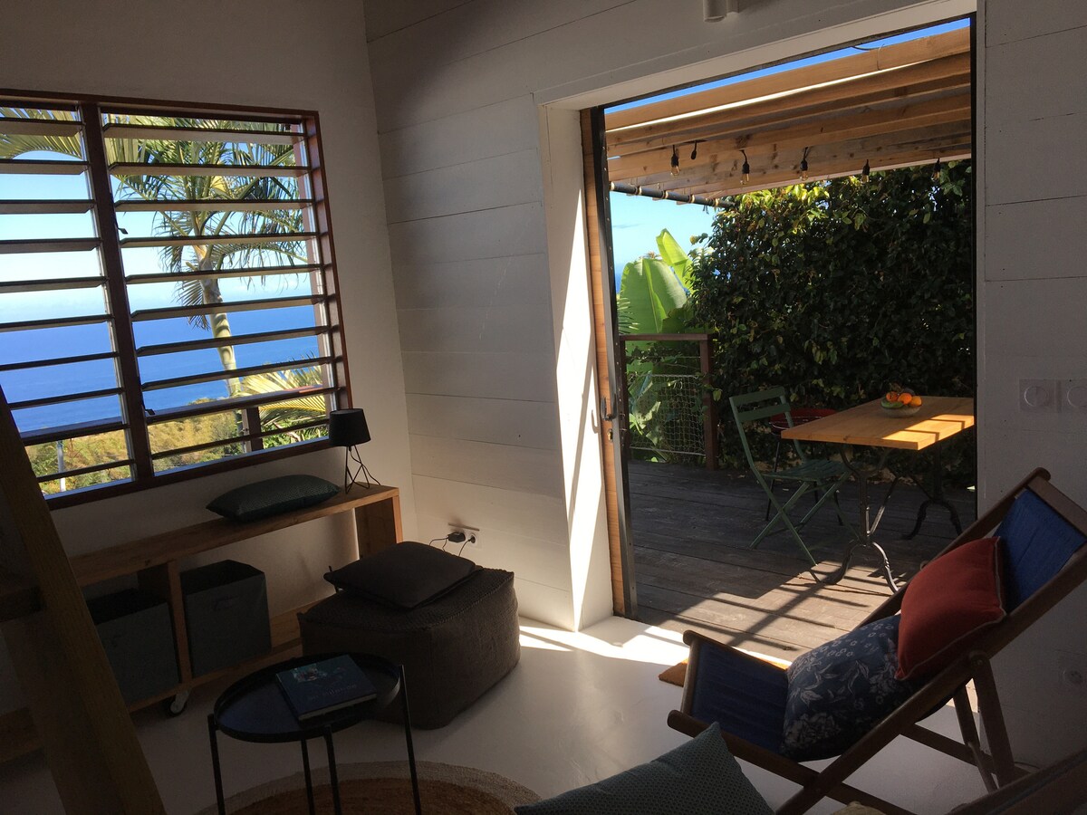 Natural light fills the living space, highlighted by a view of the ocean through slatted windows. A cozy seating area includes textured cushions and a small table. A wooden deck is visible through open doors, furnished with green chairs and a table surrounded by lush foliage.