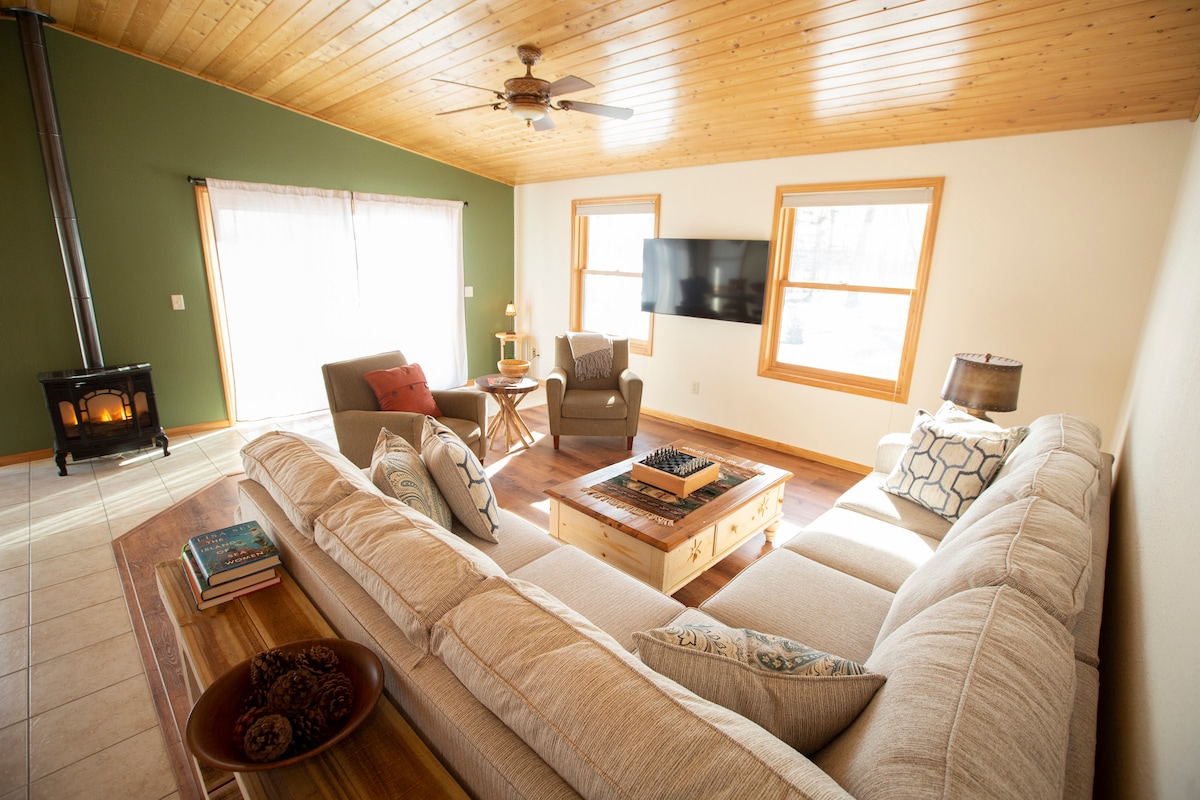 A cozy living space is depicted, featuring a light-colored sectional sofa facing a wooden coffee table and a wall-mounted television. Natural light enters through large windows, illuminating the area, while a subtle green accent wall enhances the room's calming ambiance. A small wood stove adds warmth.