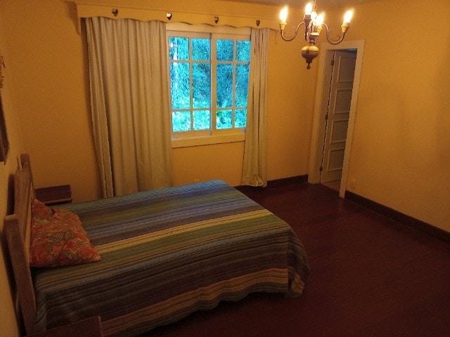 A serene bedroom is displayed, featuring a spacious bed covered with a multicolored striped blanket. Natural light filters through a large window with light curtains, contrasting with the warm yellow walls. A simple chandelier adds soft lighting to the space.