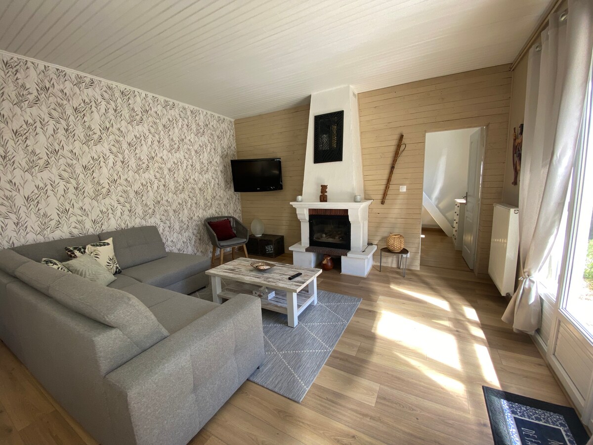 A spacious living area features a grey sectional sofa and a rustic wooden coffee table. Natural light fills the room, reflecting off the warm wooden floors. A flat-screen TV is mounted on the wall, while a decorative fireplace adds character to the inviting space.