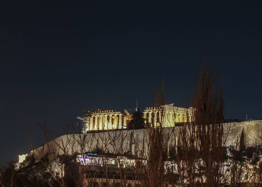 The illuminated Acropolis is prominently visible against the night sky, with its ancient columns standing tall. Silhouetted trees frame the foreground, highlighting the historic structure atop the hill. The scene conveys a sense of serenity in the heart of Athens.