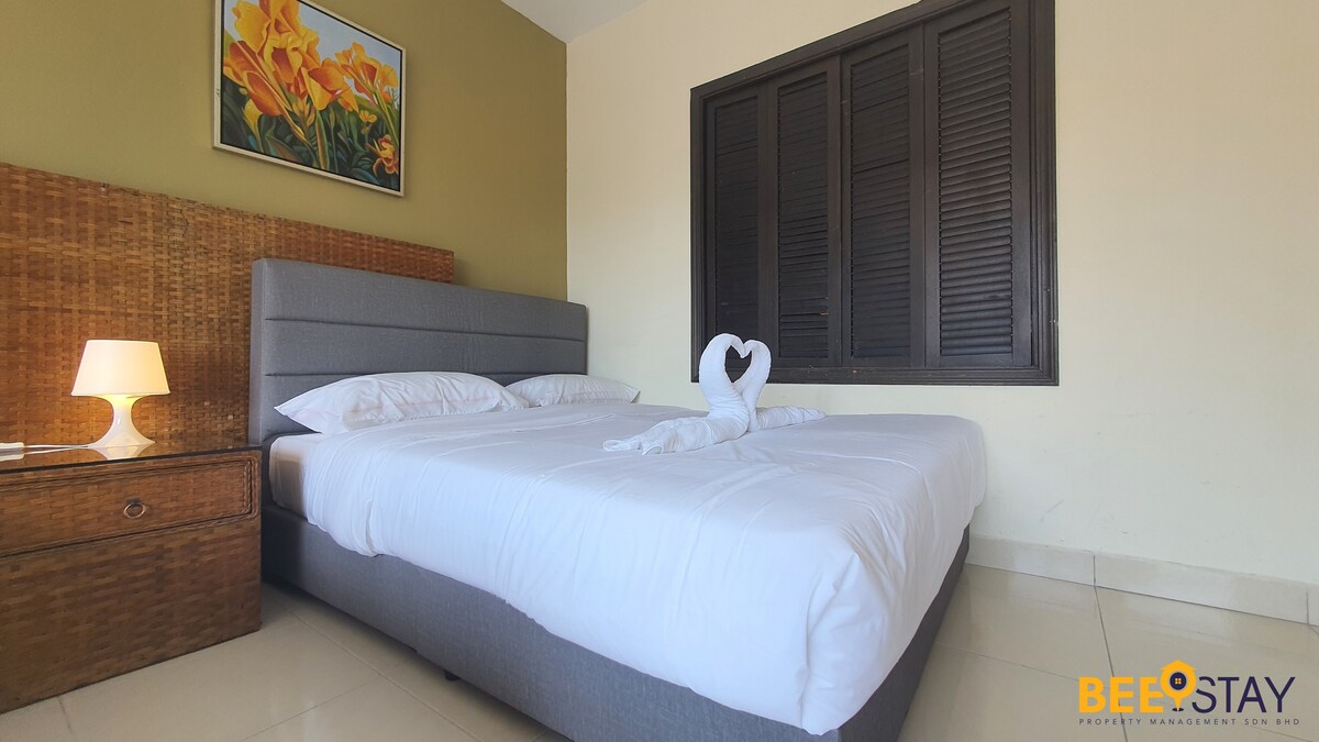 A spacious bedroom is featured, showcasing a queen bed adorned with neatly arranged linens. A decorative towel swan is positioned on the bed. The room is complemented by a bedside lamp and a woven storage unit, with a vibrant floral artwork hanging above.