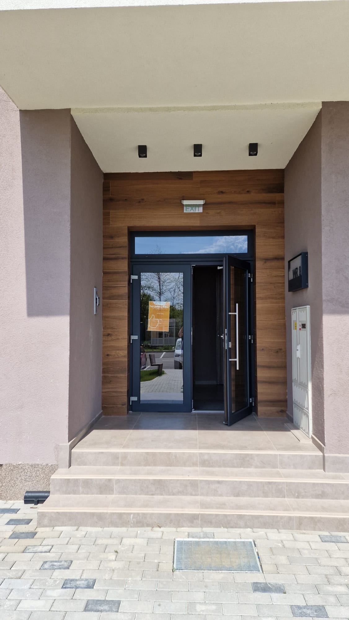 The entrance to the building is framed by modern wood paneling and features large glass doors that open outward. A tile pathway leads to the doors, while the facade is defined by neutral colored walls complemented by a minimalist light fixture.
