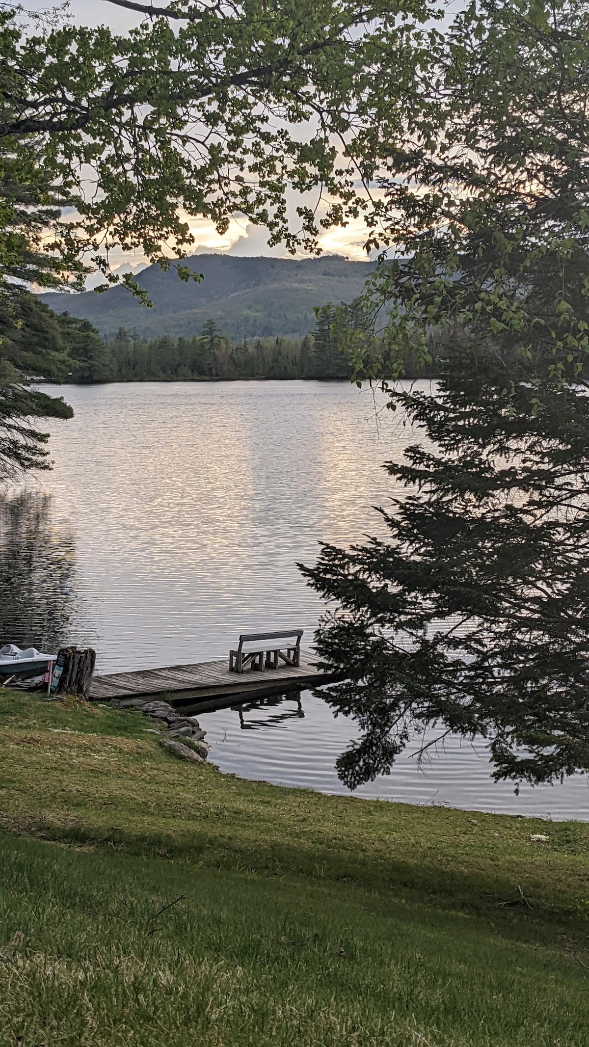The image captures a tranquil scene of Mirror Lake, with gentle ripples reflecting the clouds above. A wooden dock extends into the water, providing a serene spot for relaxation. Lush greenery surrounds the shoreline, with distant mountains creating a peaceful backdrop.