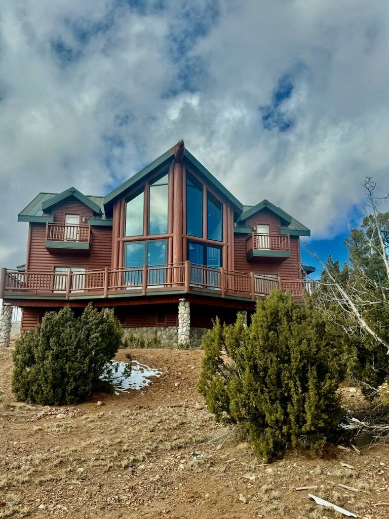 The cabin's exterior showcases a rustic design with rich wooden tones and multiple levels. Large floor-to-ceiling windows create a striking focal point on the front, surrounded by a rugged natural landscape. Stone accents at the base and a sloped roof complement the cabin's architectural style.