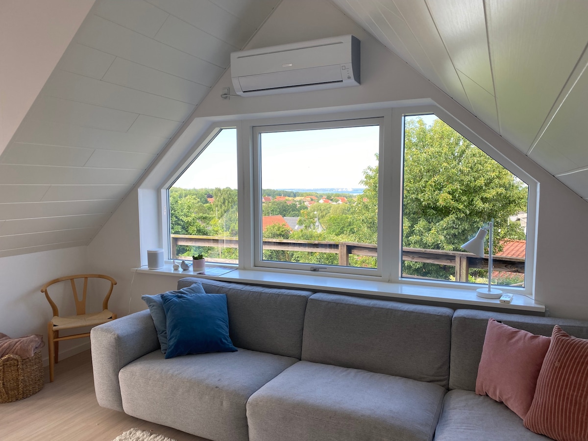 A light-filled living space features a grey sofa accessorized with a blue and a pink cushion. A rattan chair is positioned nearby, while large windows offer a view of greenery and the distant sea. The sloped ceiling enhances the cozy atmosphere.