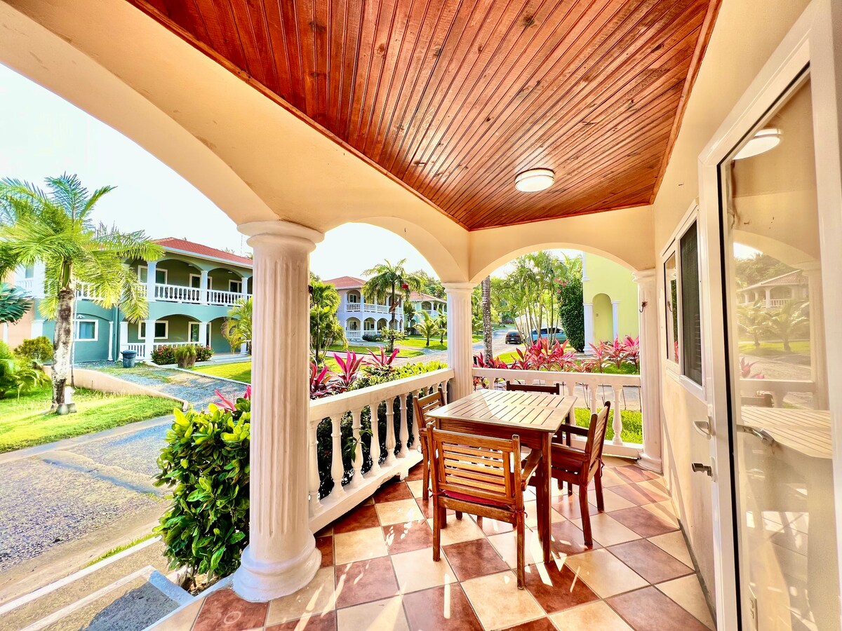 An outdoor dining area features a wooden table surrounded by matching chairs, set under a covered porch. The area is framed by decorative columns and tiled flooring. Lush greenery and colorful plants are visible in the surrounding landscape, creating a welcoming atmosphere.
