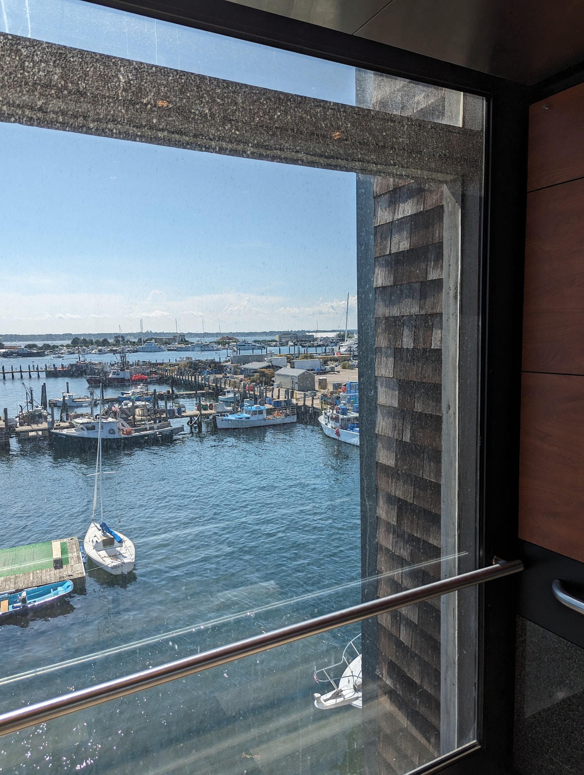 A view of a vibrant harbor is captured through a large glass window. The water reflects sunlight, while boats are moored at the docks. The scene includes docks lined with various vessels, showcasing the active maritime environment.