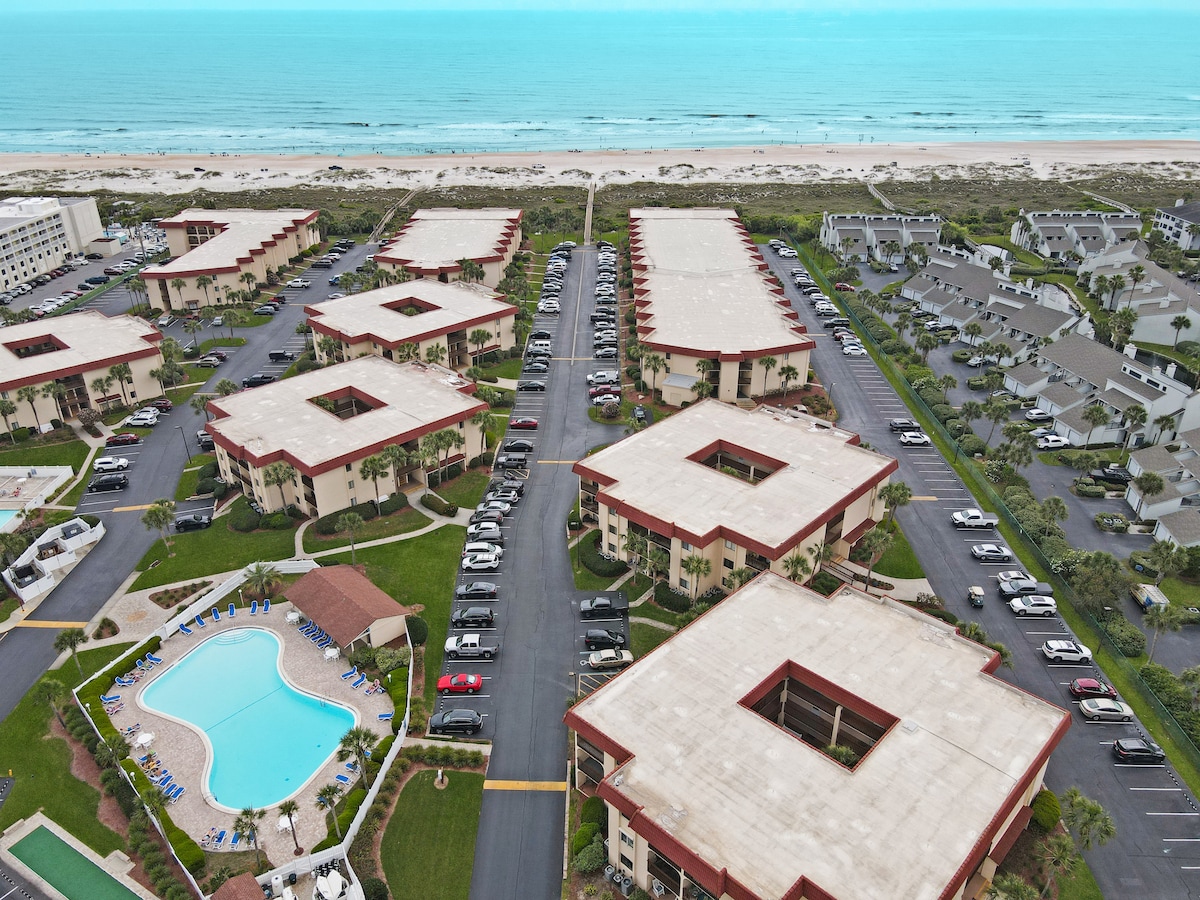 Aerial view captures multiple residential buildings arranged around a central pool area. The blue ocean water stretches along the sandy beach in the background, contrasting with the greenery of the surrounding landscape and parking areas.