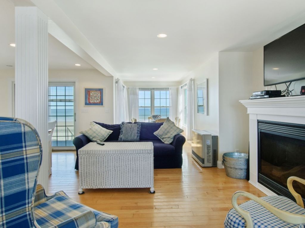 An inviting living space features large windows offering views of the beach and ocean. Comfortable seating includes a large blue sofa and patterned chairs. Light wood flooring complements the space, while a cozy fireplace adds warmth. Decor is simple and reflects a coastal theme.
