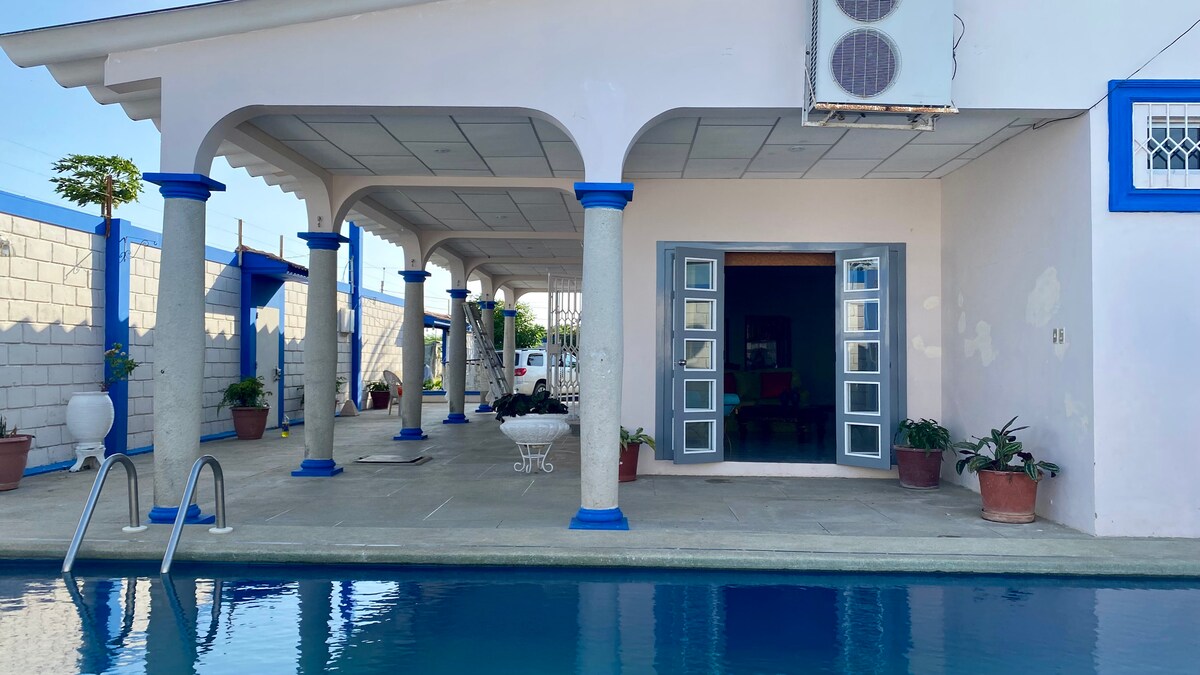 An inviting outdoor area features a swimming pool surrounded by tall columns with blue accents. Potted plants are placed around the pool, and large double doors open into a spacious interior, enhancing the connection between indoor and outdoor spaces.