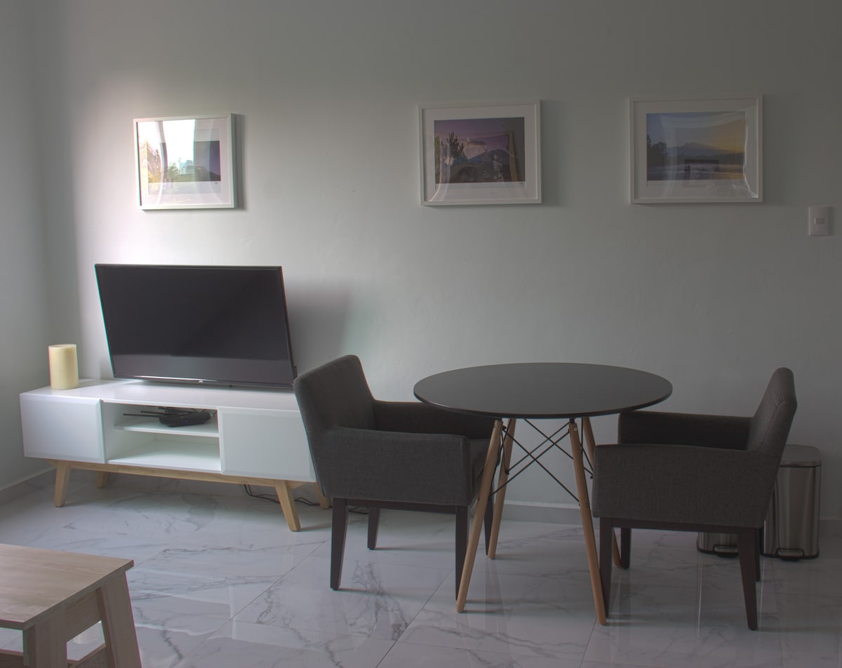 A minimalist living area is presented with a round dining table and two gray armchairs. A television is positioned on a white media console against the wall, which is adorned with framed artwork. Natural light brightens the space, highlighting the marble flooring.