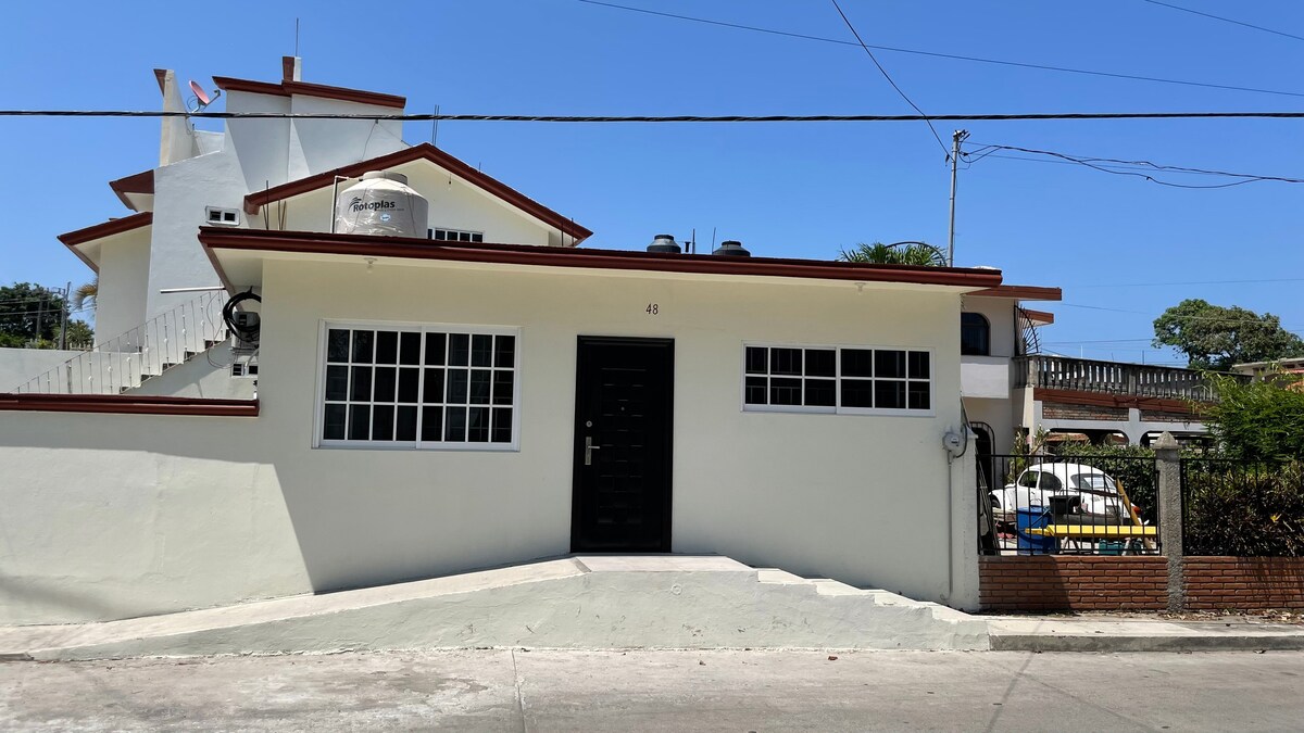 The exterior of the accommodation is shown, featuring a single-story white facade with large windows allowing natural light. A black front door welcomes guests, and the property is set against a clear blue sky. A gentle slope leads to the entrance, providing easy access.