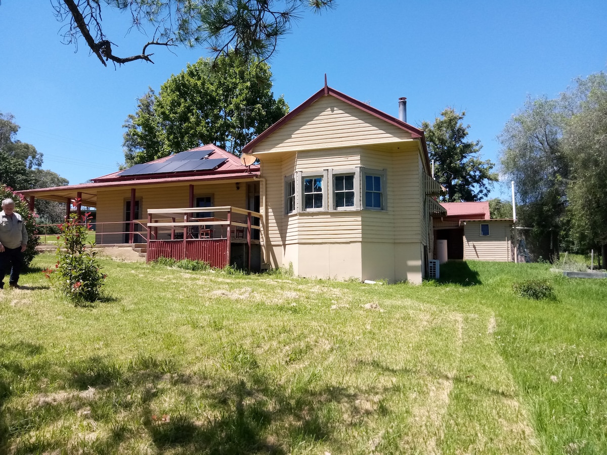 The exterior of a spacious farmhouse showcases a traditional design with a front porch and multiple windows. A lush green lawn surrounds the house, while trees provide natural shade. The building features a red roof and light-colored walls, suggesting a harmonious blend with the landscape.