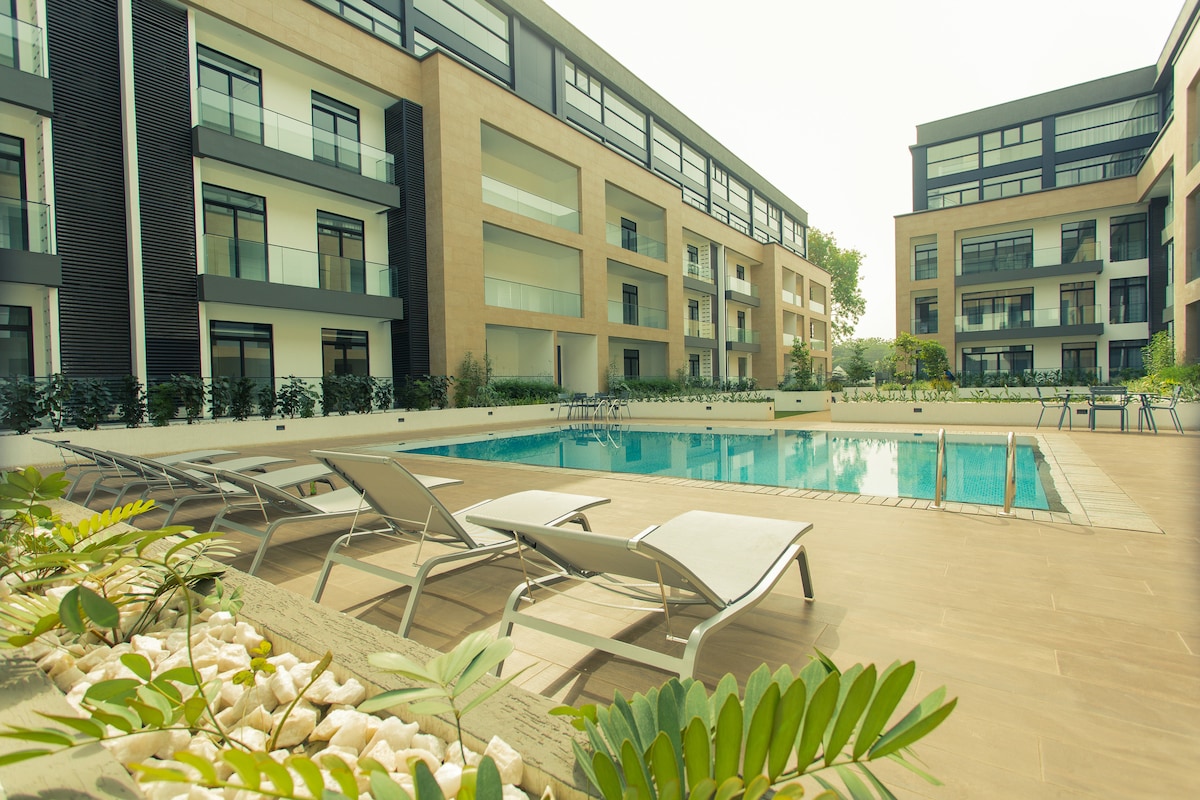 An outdoor pool area is highlighted, surrounded by modern apartment buildings with large windows. Chaise lounges are positioned along the poolside, offering a place to relax. Lush greenery and decorative stones enhance the area, contributing to a serene outdoor ambiance.