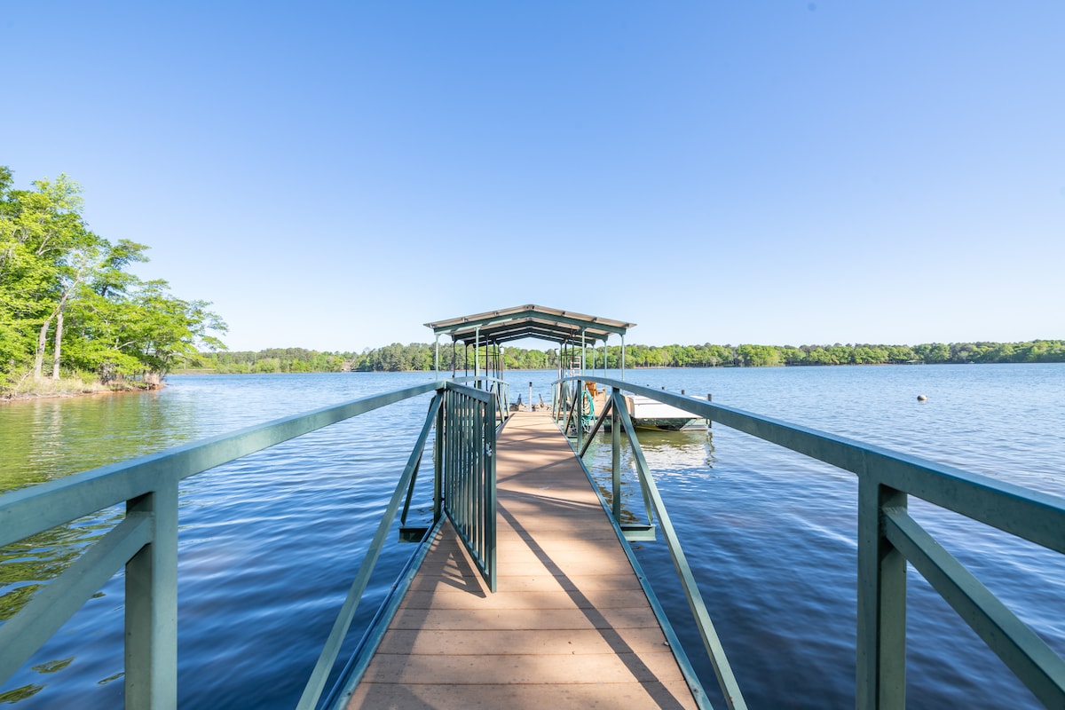 A spacious wooden dock extends over calm waters, providing access to a shaded area covered by a roof. Lush greenery borders the lake, enhancing the serene surroundings, with clear blue skies overhead reflecting off the water.