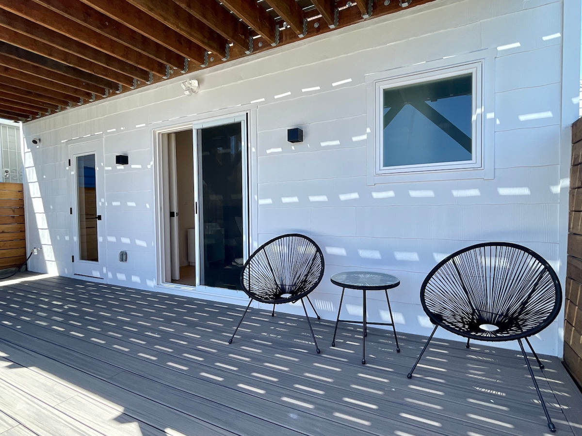 A spacious deck features two black, modern chairs and a small round table, surrounded by horizontal slats that create a pattern of light and shadow. The deck provides direct access to the indoor living area through sliding glass doors.
