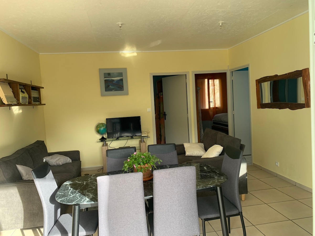 A light-filled living area is furnished with a gray sofa and a dark glass dining table surrounded by several chairs. A mounted shelf displays decorative items, while a television is positioned nearby. Doorways lead to separate rooms, with soft tones enhancing the space.