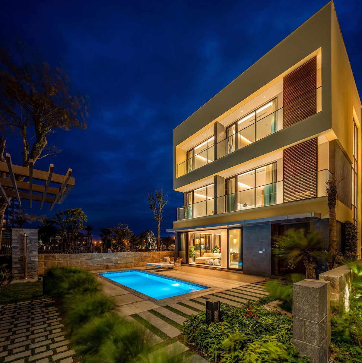 The modern villa is illuminated at night, showcasing large glass windows and a private pool in the foreground. The outdoor area features lush landscaping and a spacious patio, creating an inviting setting for relaxation and gatherings.