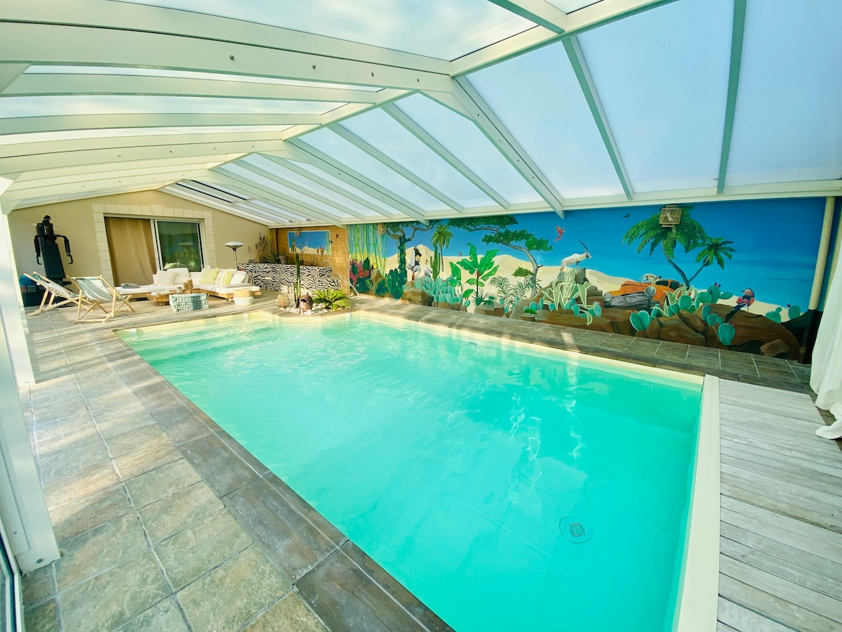 An indoor pool area features a bright turquoise pool beneath a clear, sloped roof, allowing for ample natural light. A vibrant mural adorns one wall, depicting a tropical scene. Lounge chairs and a small table are arranged nearby, promoting relaxation.