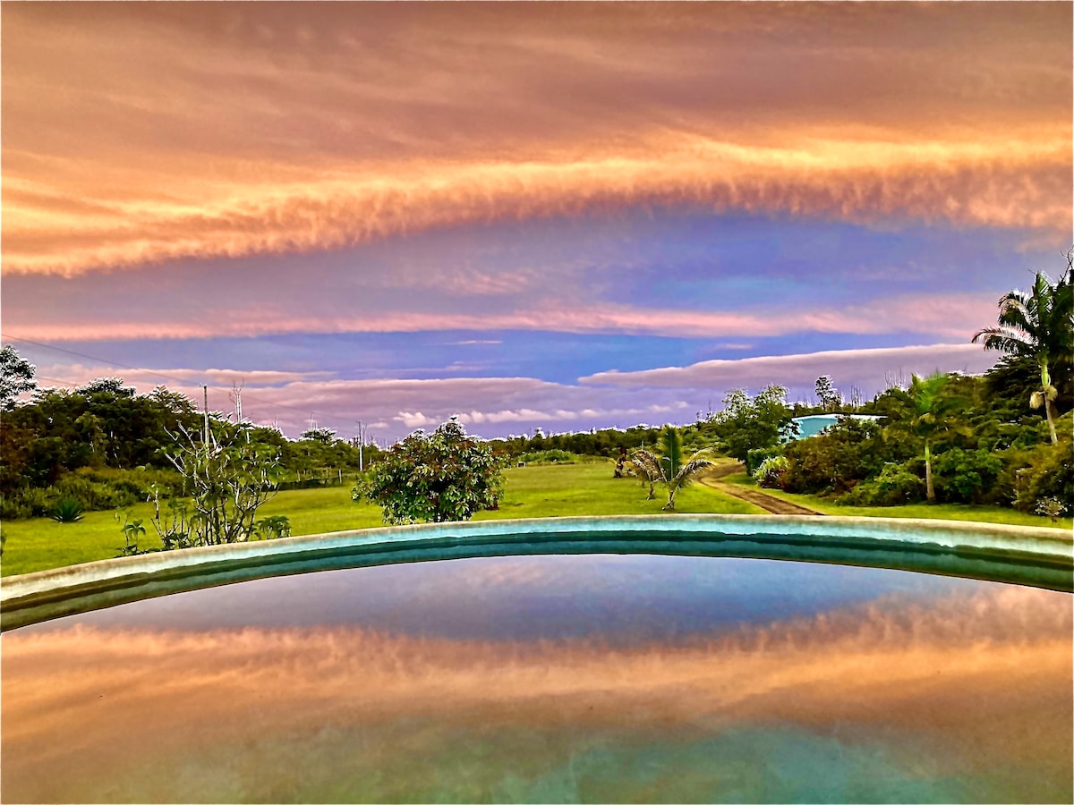 The image captures a serene view of lush greenery under a colorful sunset sky. The tranquil reflection of clouds in a nearby plunge pool adds depth to the landscape, highlighting the peaceful surroundings of the retreat.