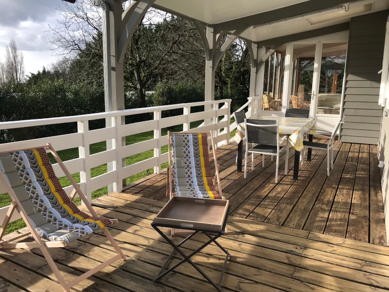 A spacious outdoor terrace features wooden decking with a dining table and chairs. Two lounge chairs, adorned with colorful patterns, are positioned for relaxation. The surrounding greenery enhances the tranquil setting, with a partial view of the garden beyond.