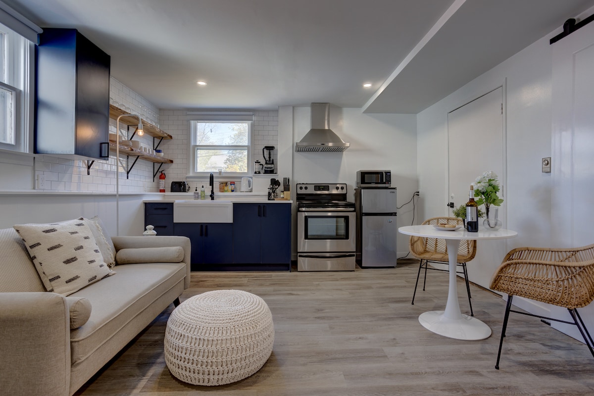 A modern kitchen is equipped with stainless steel appliances, including a stove and microwave. Open shelving displays kitchen essentials. A cozy seating area features a light-colored sofa and a round dining table with woven chairs, creating a functional and inviting space.