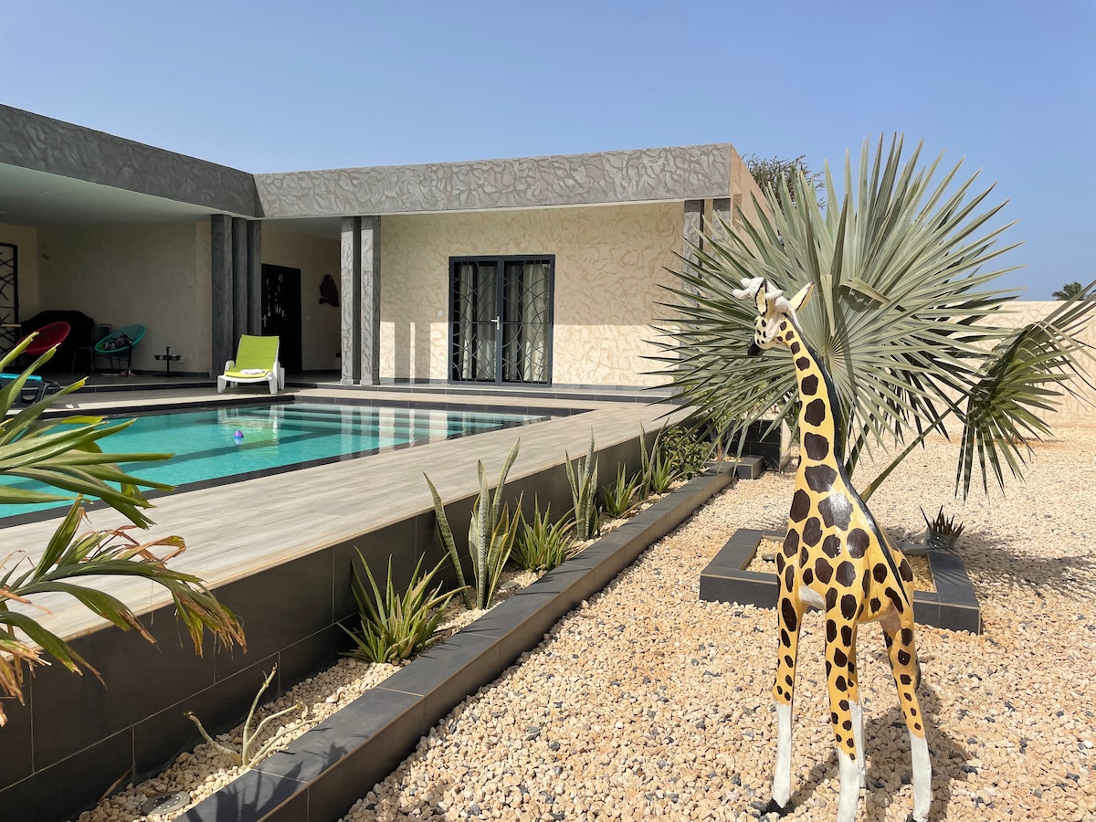 An outdoor area is depicted with a vibrant pool surrounded by tropical plants and a decorative giraffe statue. Lounge chairs in bright green are positioned near the pool, alongside a patio overlooking the serene landscape.