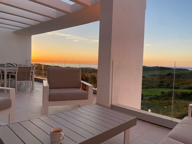 An outdoor terrace features modern furniture, including a comfortable sofa and chairs arranged around a sleek coffee table. A stunning sunset illuminates the sea view, blending warm colors on the horizon. The terrace is shielded by glass railings, enhancing the peaceful ambiance.