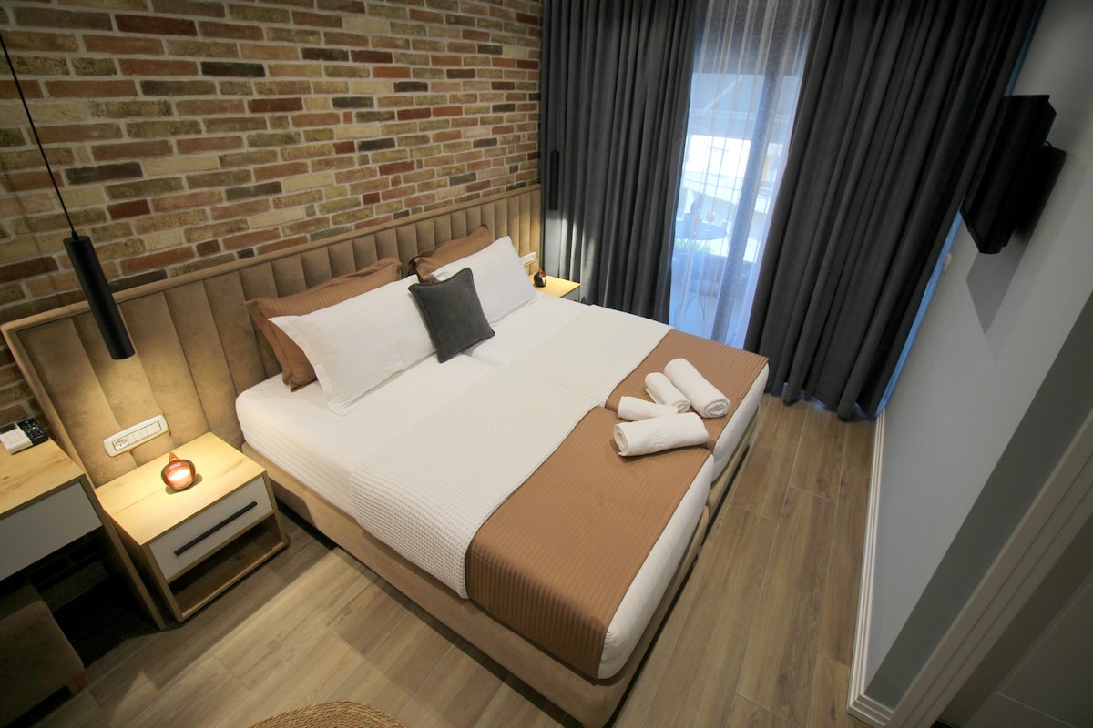 A cozy bedroom features a spacious bed with a neutral-toned blanket and decorative pillows. A brick wall adds texture, while dark curtains frame a window, allowing light to filter in. A bedside table with a lamp complements the warm, calming environment.