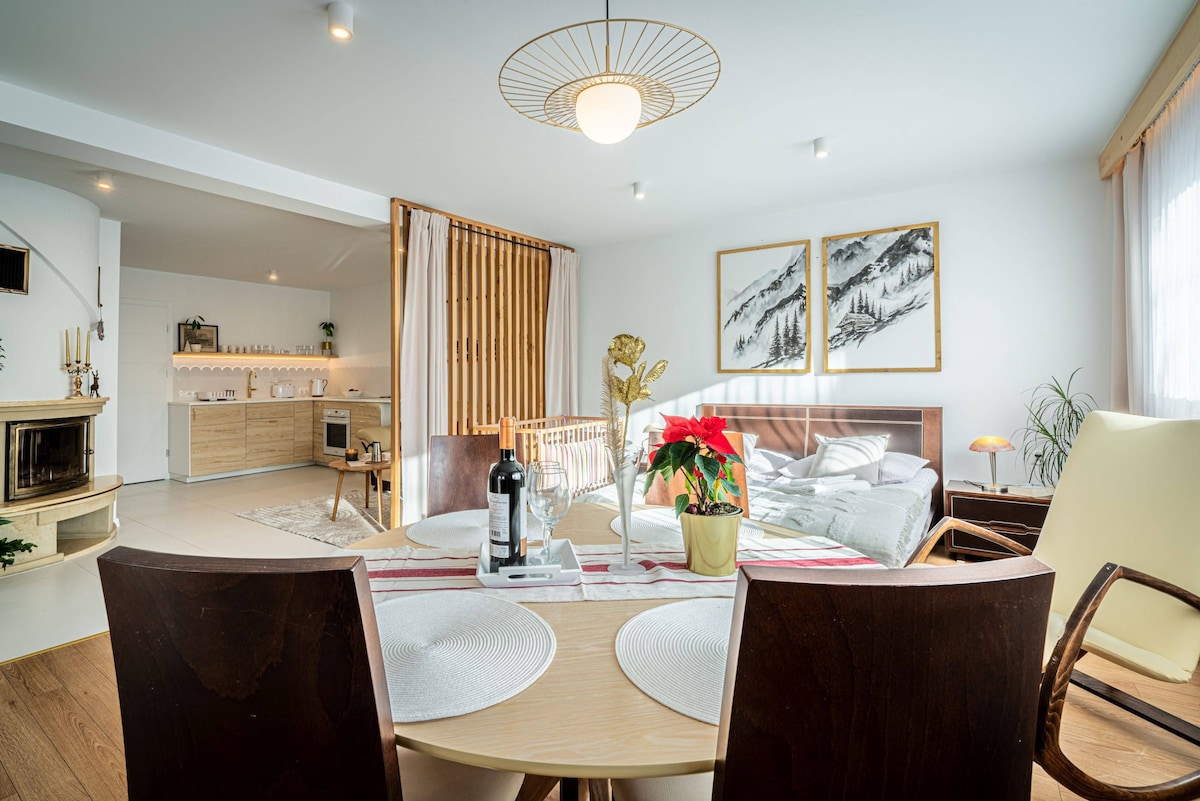 A dining area is presented with a round table set for two, featuring placemats and a decorative plant. The living space is visible in the background, showcasing a cozy lounge with a spacious bed, inviting light, and a modern kitchen area with wooden accents.