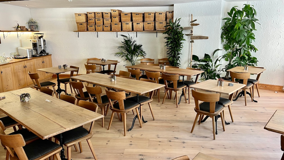 An inviting communal dining area features wooden tables paired with comfortable chairs, arranged to encourage social interaction. Natural light filters through the space, highlighting potted plants and woven storage baskets along the wall, contributing to a warm, welcoming ambiance.