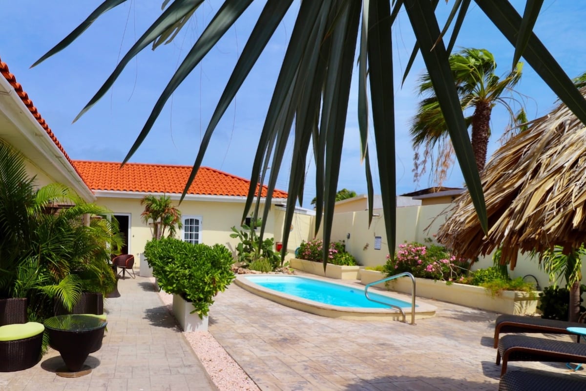 A shared pool is surrounded by lush greenery and tropical plants. Comfortable outdoor seating areas are positioned nearby, and a thatched-roof structure offers shade. The pool area is complemented by a tiled patio and features a light color palette, creating a relaxed outdoor environment.