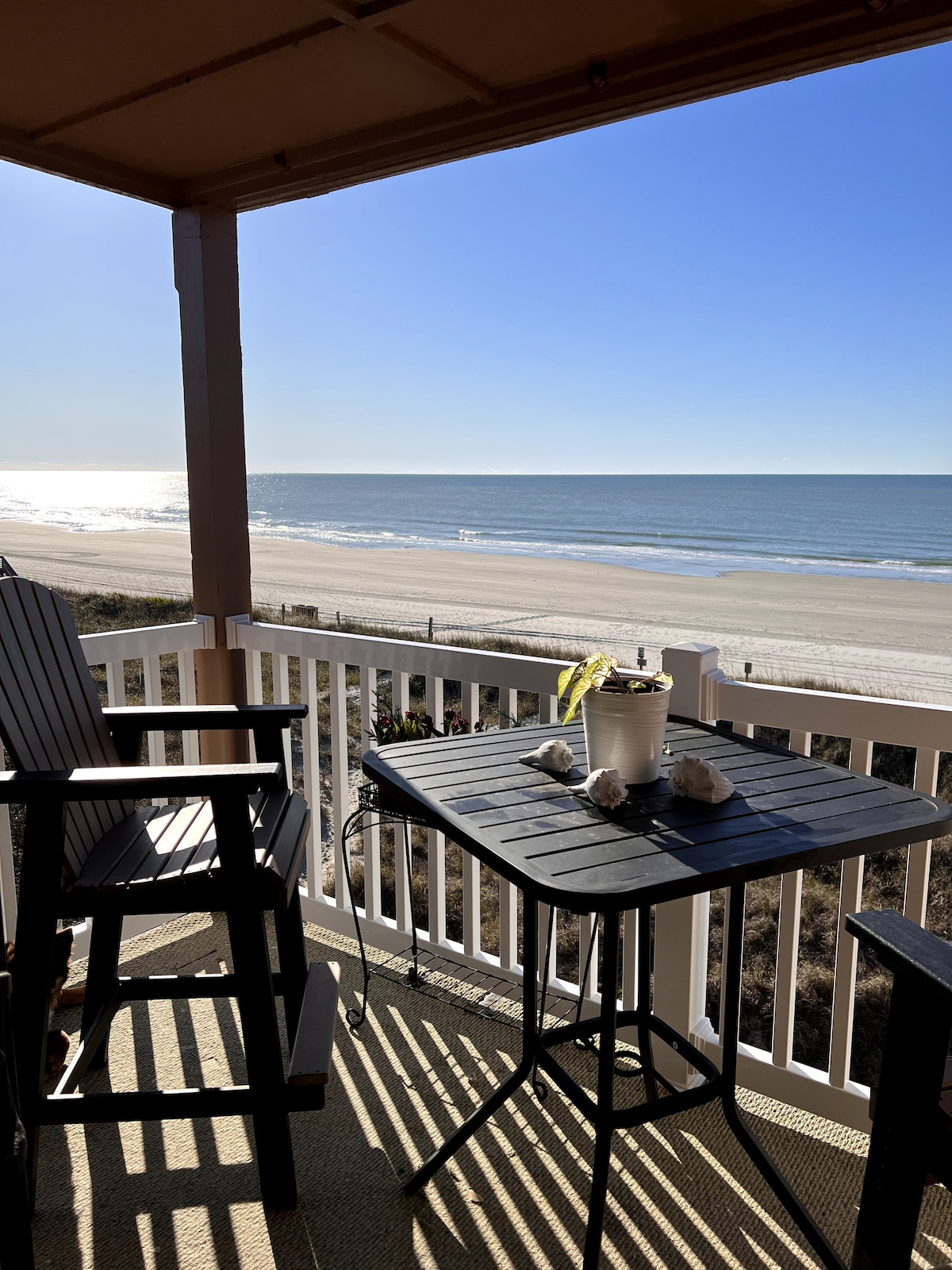 A balcony view showcases the ocean and sandy beach under a clear sky. A small table is set with a potted plant and seashells, while two rocking chairs provide seating. Sunlight casts gentle shadows, enhancing the relaxing atmosphere of the space.