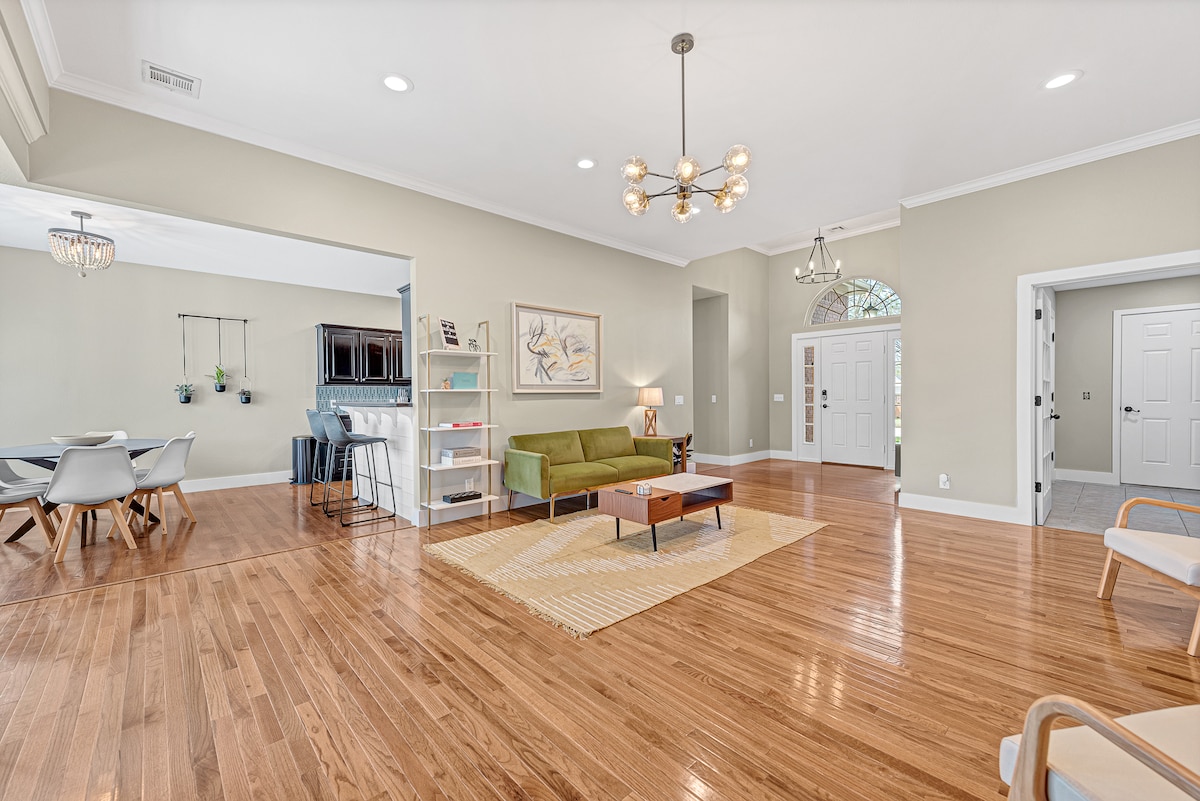 A spacious living area features warm wooden flooring and is illuminated by natural light. A modern green sofa and a low coffee table are positioned at the center. The surrounding walls are painted in neutral tones, and a stylish chandelier adds a contemporary touch.