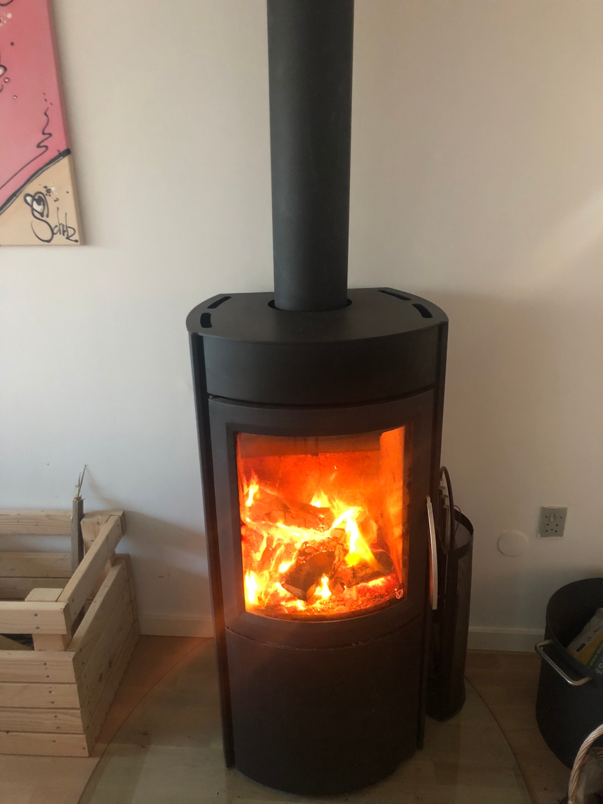 A modern wood-burning stove stands against a neutral wall, showcasing a vivid display of flames through its glass front. A wood storage bin is placed beside it, enhancing the warmth and comfort of the space.