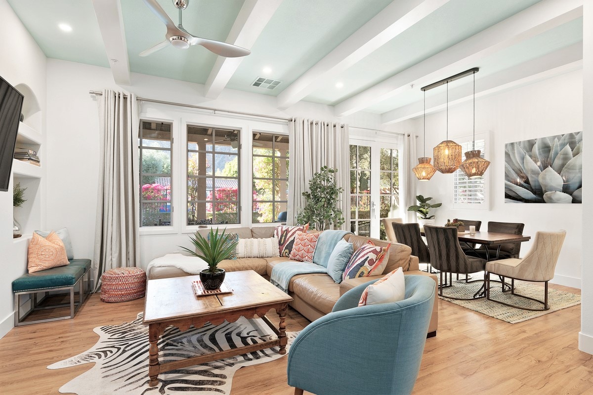 A spacious living area is illuminated by natural light through large windows. A comfortable sectional sofa is adorned with colorful pillows, positioned on a patterned area rug. A wooden coffee table sits center stage, while a dining area with a dark table and chairs is visible in the background.