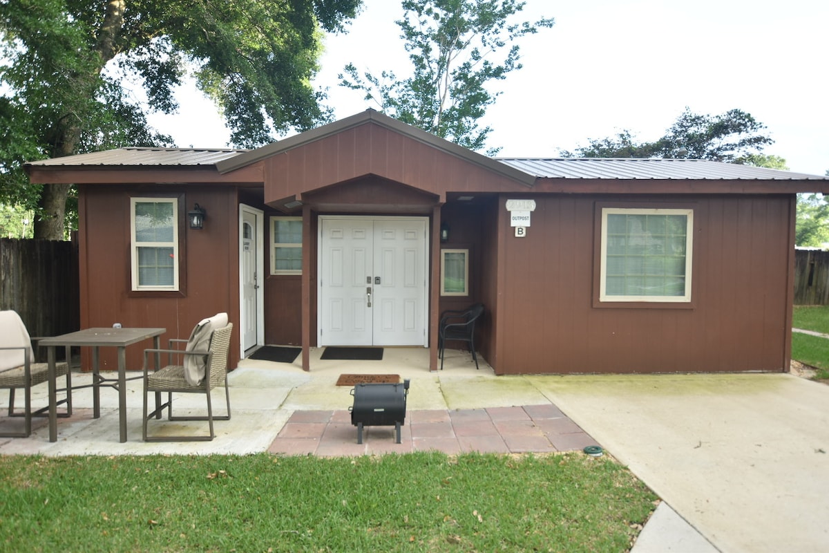 The exterior of the building features a welcoming brown facade with two front doors. A small, circular table with four chairs is positioned on a patio area. A charcoal grill rests on the ground nearby, surrounded by a grassy yard and shaded by trees.