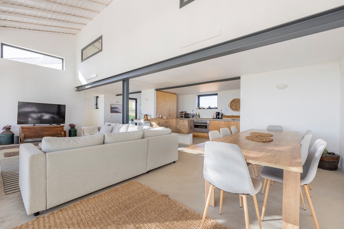 A spacious living area features a light-colored sectional sofa paired with a wooden dining table surrounded by grey chairs. Large windows allow natural light to fill the space. The kitchen area is visible in the background, showcasing modern appliances and wooden cabinetry.