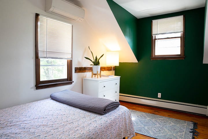 This cozy bedroom features a comfortable bed, ample natural light, and a touch of greenery. The serene green accent wall and simple decor create a peaceful atmosphere, perfect for a restful night's sleep. Ideal for relaxation and comfort.