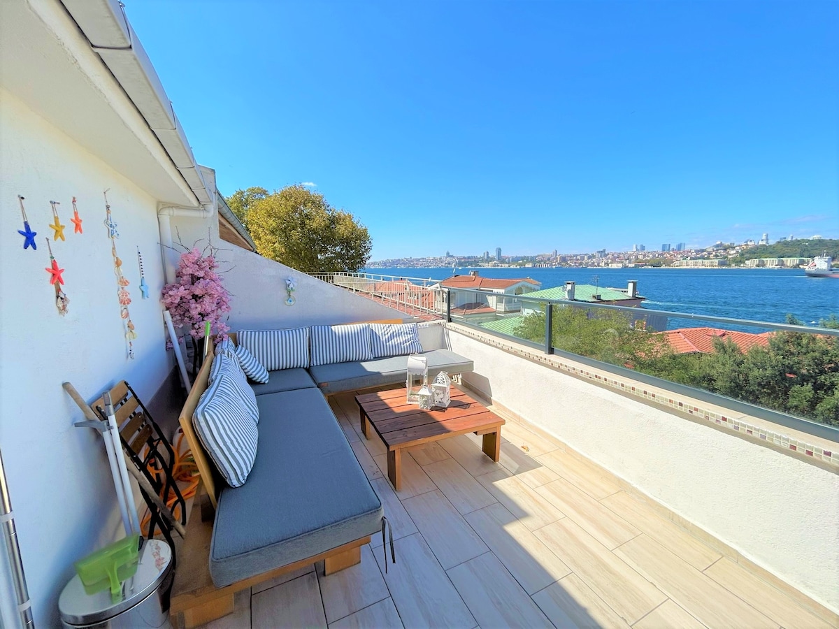 A spacious terrace provides a comfortable seating area with cushioned benches and a wooden coffee table. The area offers panoramic views of the water and cityscape, framed by a clear blue sky. Decorative elements like wall art add a personal touch.