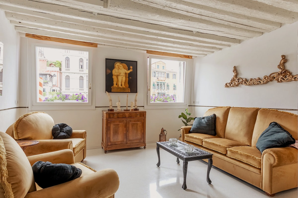 A bright living area is showcased with large windows allowing natural light to fill the space. Two comfortable sofas, accented with pillows, face a wooden coffee table. Decorative art pieces are mounted on the walls, creating an elegant touch, while potted plants enhance the inviting atmosphere.