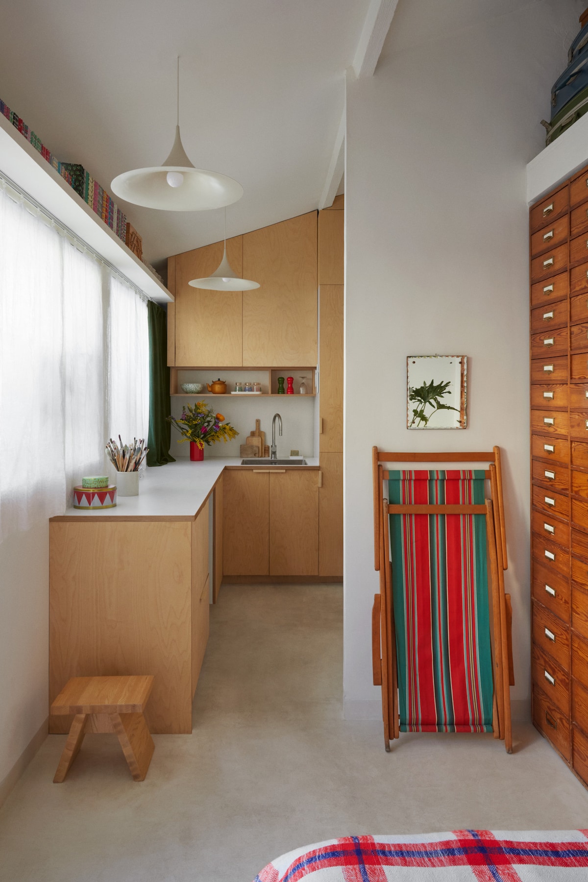 A compact kitchen area features wooden cabinetry, with a countertop providing workspace. Natural light filters in through the large window, illuminating a vase of flowers on the sink. A colorful, striped chair is positioned next to a wooden stool, enhancing the space's warmth and functionality.