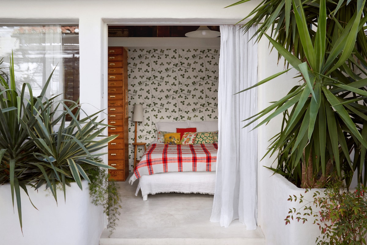A bright sleeping area is framed by large glass doors and adorned with a patterned wallpaper. A spacious bed is dressed in bright linens, complemented by colorful pillows. Lush green plants flank the entrance, enhancing the inviting ambiance.
