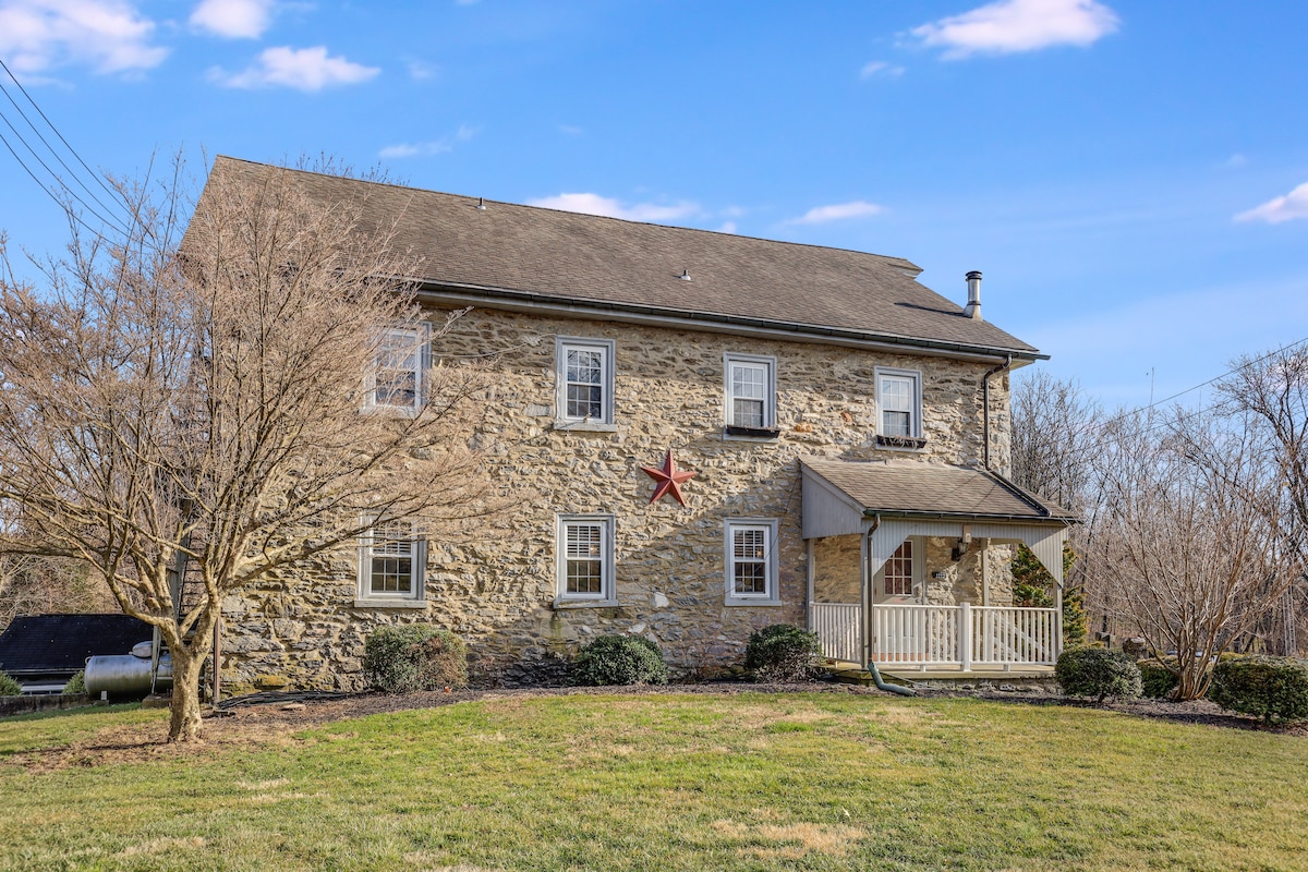 The exterior of a charming stone building is depicted, featuring multiple large windows and a welcoming front porch. A red star adorns the facade, enhancing the rustic appeal. Lush green grass surrounds the structure, contributing to the serene countryside atmosphere.
