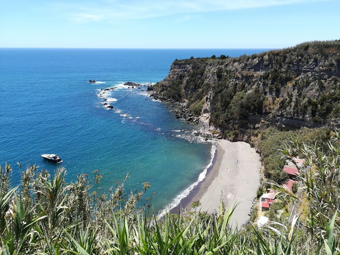 A serene coastal view is presented, showcasing a secluded beach framed by cliffs and vegetation. The clear blue waters reflect the sky, while a small boat can be seen near the shoreline. Gentle waves lap at the sandy beach, enhancing the tranquil scene.
