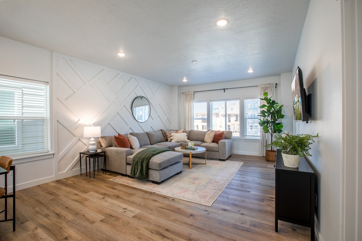 The living area features a spacious sectional sofa arranged around a coffee table, complemented by soft, warm lighting. A large window invites natural light and offers views of the outside, while a decorative plant adds a touch of greenery to the space.